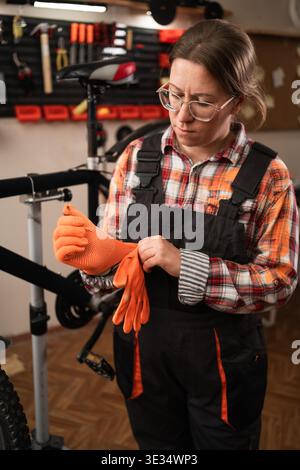 Female mechanic in glasses putting on orange gloves and preparing for repair work in authentic bicycle shop. Concept of professional repair and craft. Stock Photo