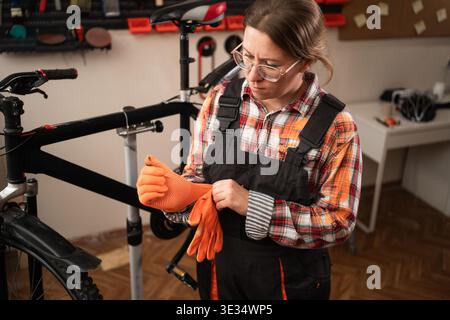 Female mechanic in glasses putting on orange gloves and preparing for repair work in authentic bicycle shop. Concept of professional repair and craft. Stock Photo