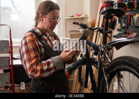 Woman bike mechanic using laptop for online diagnostics in workshop. Female technician searching for bicycle repair solution on internet. Concept of t Stock Photo