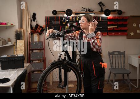 Young woman listening to a voice message on her phone while repairing bicycle in workshop, adjusting wheel mechanism. profession, technology and peopl Stock Photo