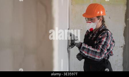 Female worker in hardhat polish wall with sponge trowel for smoothing plaster surface, copy space Stock Photo