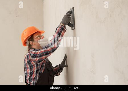 Builder woman in hardhat and protective mask sanding and smoothing walls after plastering for leveling getting it ready for painting during home renov Stock Photo