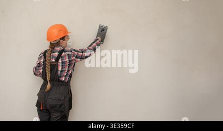 Builder woman in hardhat and protective mask sanding and smoothing walls after plastering for leveling getting it ready for painting during home renov Stock Photo