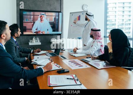 Multiethnic executives in a modern dubai office collaborate on strategy, with a colleague presenting data on a whiteboard and another joining via larg Stock Photo