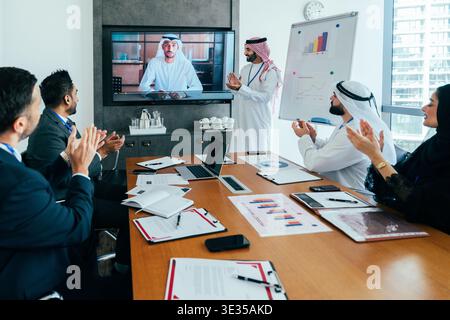Diverse business professionals attending a corporate meeting in a modern office, clapping for a presentation displayed on a large screen with a speake Stock Photo