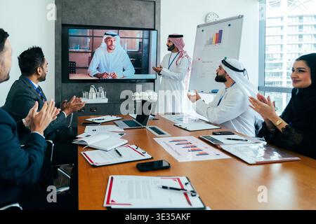 Multiethnic business people in a modern dubai office attending a video conference, applauding a virtual participant during an important corporate pres Stock Photo