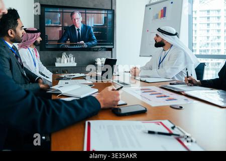 Multiethnic business people participating in a corporate board meeting, collaborating remotely with a virtual participant on a large screen in a moder Stock Photo