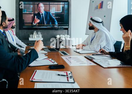 Diverse corporate team members, including arab individuals, attending a hybrid business meeting in a modern office, engaged in a virtual discussion wi Stock Photo