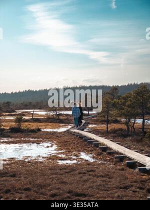 Two hikers walking wooden boardwalk over Kakerdaja bog with frozen pools and forest backdrop, Estonia Stock Photo