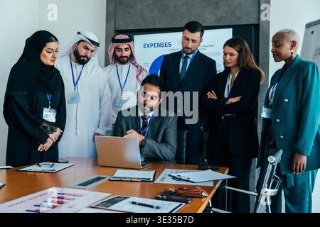 Multiethnic business people gathered around a laptop in a modern conference room, attentively discussing financial data and expenses presented on a sc Stock Photo