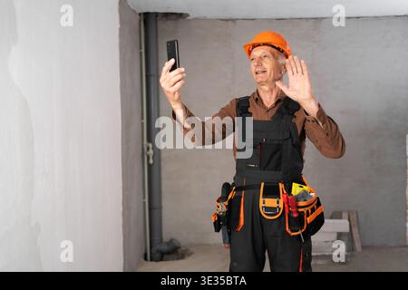 Senior builder in hardhat making video call using smartphone at construction site. building and technology concept Stock Photo