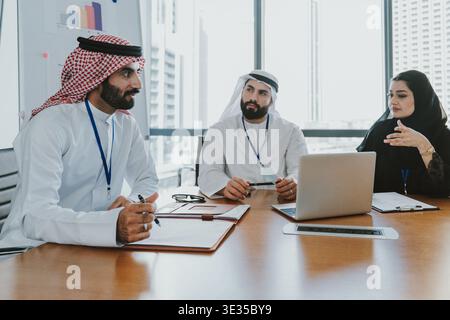 Three middle eastern business professionals, two men in traditional thobes and a woman in an abaya, collaborating around a desk in a modern office dur Stock Photo