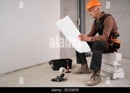 senior contractor in helmet and overalls reading paper blueprint showcasing expertise in construction industry. profession, construction and repair co Stock Photo