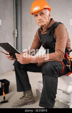 Portrait of pensive builder or repairman senior man in safety helmet on building site during renovation process. profession, construction and repair c Stock Photo