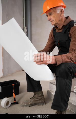 senior contractor in helmet and overalls reading paper blueprint showcasing expertise in construction industry. profession, construction and repair co Stock Photo