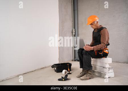 builder or repairman senior man in safety helmet sitting using laptop computer ordering online on building site during renovation process. profession, Stock Photo