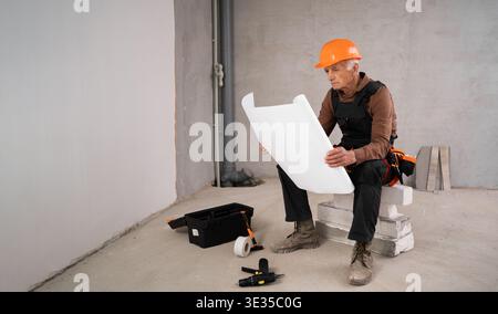senior contractor in helmet and overalls reading paper blueprint showcasing expertise in construction industry. profession, construction and repair co Stock Photo