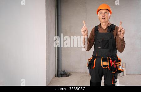 Senior repairman or construction worker in safety helmet on building site, sitting near a toolbox, pointing index fingers up at an ad placement area. Stock Photo
