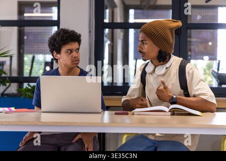 Diverse male students sitting at long wooden table studying with laptop, textbook and headphones Stock Photo