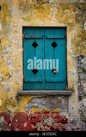 Vertical composition of rustic turquoise wooden window with black diamond details on distressed yellow, white and red painted wall, Mediterranean arch Stock Photo