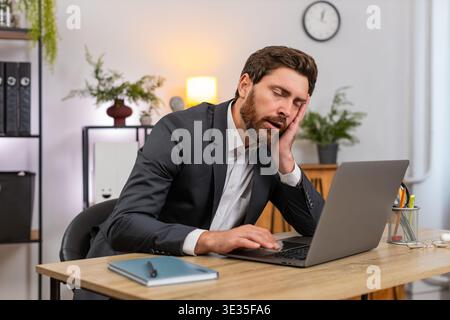 Middle-aged businessman at home office types report on laptop gets sleepy and nods off leaning on hand. Freelancer guy at table fights fatigue blinks slow and almost sleeps then returns to work again. Stock Photo