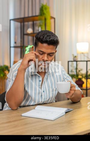 Indian man at home drinks coffee and writes goals in notebook ticks boxes and plans day with drive. Arabian guy at desk reviews list adds tasks sets priorities smiles feels focus and motivation aim Stock Photo