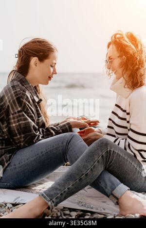 Beach friends dice two happy women in jeans sit on a blanket playing games together by the ocean waves Stock Photo