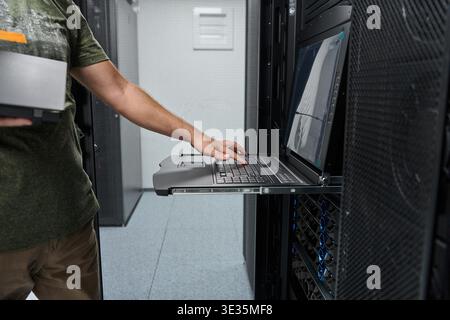 IT technician using a rack-mounted laptop console to maintain servers in a data center, monitoring hardware and performing configuration and diagnostics on networked equipment behind a perforated cabinet door. Stock Photo