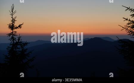 Sun sets behind distant mountains, casting soft orange and pink glow across sky. Silhouetted pine trees frame foreground, while layers of dark hills create serene and tranquil scene. Stock Photo