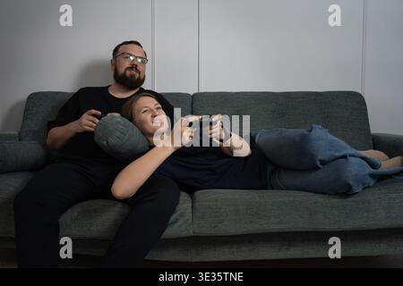 Young couple enjoying their leisure time at home, a woman lying on a man's lap while both are focused on playing a video game using wireless controlle Stock Photo