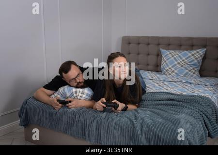 Young man and woman lying on a comfortable bed, engrossed in a competitive gaming session with controllers, enjoying their hobby in a domestic setting Stock Photo