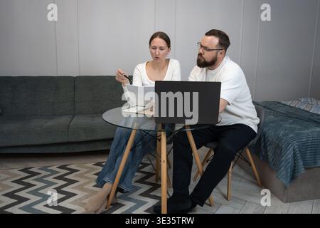 Young adult couple collaborating and sharing ideas, analyzing financial papers and using a laptop while sitting at a glass table in their modern home Stock Photo