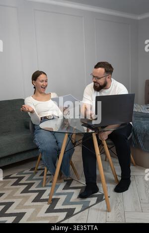 Happy couple smiling and discussing important documents while working from home, sharing a laptop and celebrating success on a financial or project re Stock Photo