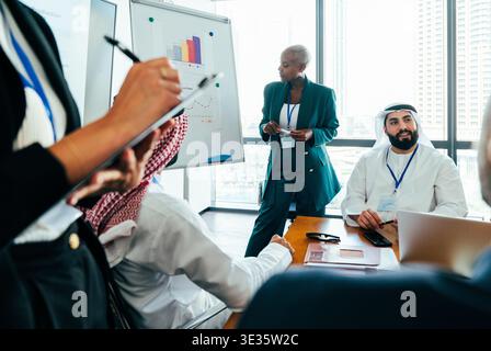 Diverse group of professionals discussing project data, with a woman presenting charts on a whiteboard to arab businessmen and other colleagues in a m Stock Photo