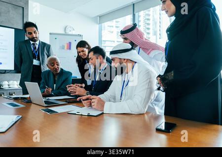 Diverse multinational executives collaborating around a laptop in a modern dubai boardroom, analyzing data and strategizing for global growth, partner Stock Photo