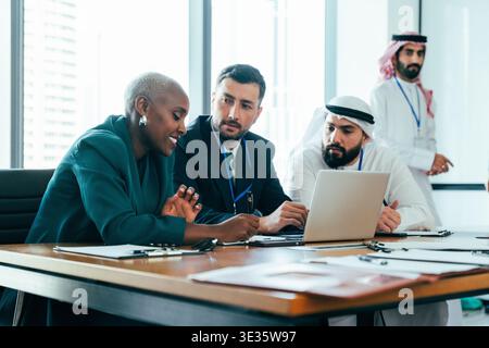 Multiethnic business professionals are discussing project details, working together at a conference table, using a laptop, and collaborating in a mode Stock Photo