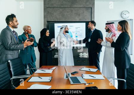International business team in a dubai office applauding as two partners shake hands, celebrating a successful multicultural corporate agreement and p Stock Photo