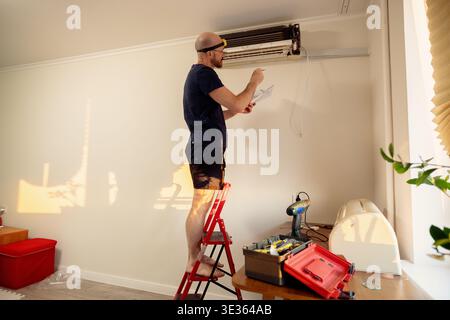 A man stands on a red ladder while inspecting and repairing an open wall-mounted air conditioner unit in a living room with warm evening sunlight. Stock Photo