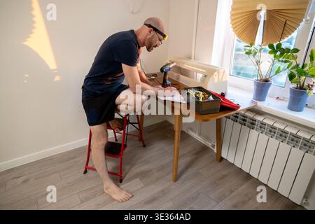 A focused man in a headlamp and glasses sits at a wooden table, writing notes and planning a home improvement project with tools spread around him. Stock Photo