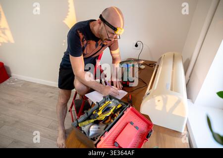 A focused man with a headlamp sits on a ladder, taking notes while repairing an air conditioning indoor unit on a desk surrounded by tools. Stock Photo