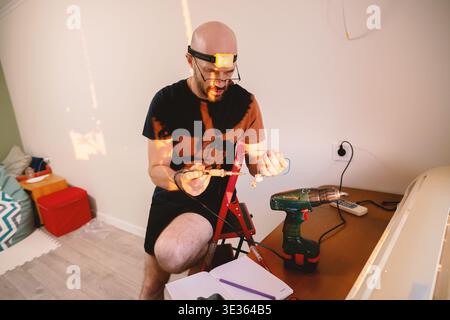 A concentrated man performs precision soldering on electronic components at his desk, using a headlamp for better visibility during a home improvement Stock Photo