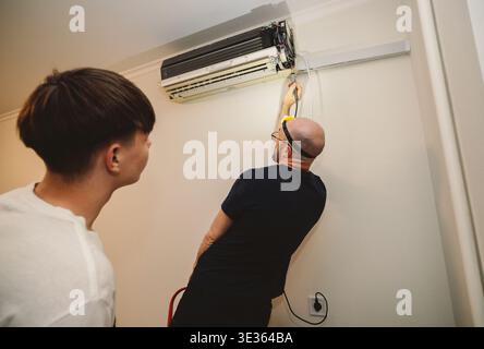 A skilled service man using a headlamp performs maintenance on a split-system air conditioner while a young man observes the process indoors. Stock Photo