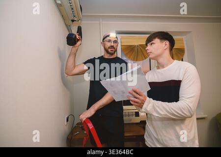An adult technician and a teenager collaborate on a home improvement project, installing an indoor air conditioning unit using a drill and reading a m Stock Photo