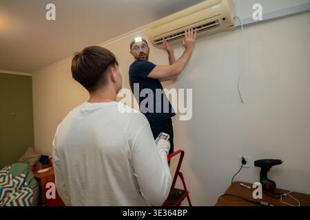 A professional technician with a headlamp performs maintenance on a wall-mounted air conditioning unit in a domestic apartment while the homeowner loo Stock Photo