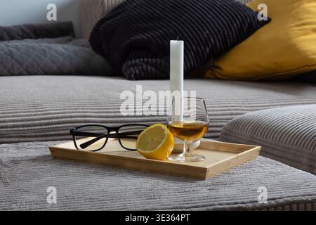 Whiskey glass, lemon and eyeglasses placed on a wooden tray with a white candle on a textured sofa. Soft ambient light creates a calm, intimate living Stock Photo