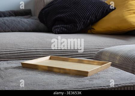 An empty wooden tray rests on a textured grey sofa, surrounded by soft pillows in navy and mustard, creating a cozy and inviting scene Stock Photo