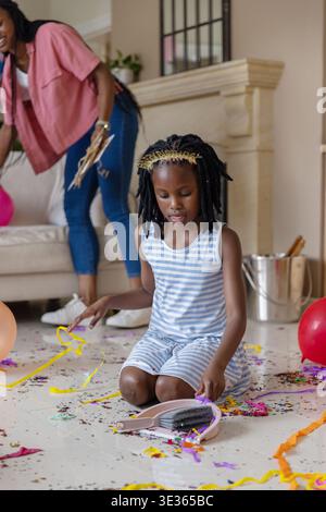African American mom and child kneeling, picking confetti with dustpan in living room, balloons Stock Photo