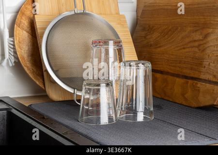 Clear drinking glasses placed upside down on a drying mat near wooden cutting boards and a metal sieve. Bright kitchen scene highlights cleanliness, o Stock Photo