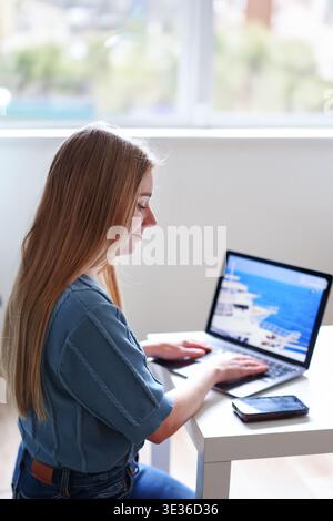 Female freelancer using laptop and smartphone at home planning vacation online. Remote work and travel concept. Stock Photo
