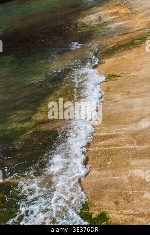 Gentle Wave Lapping Against Rocky Shore With Sea Foam and Algae on Sunlit Coastline Stock Photo